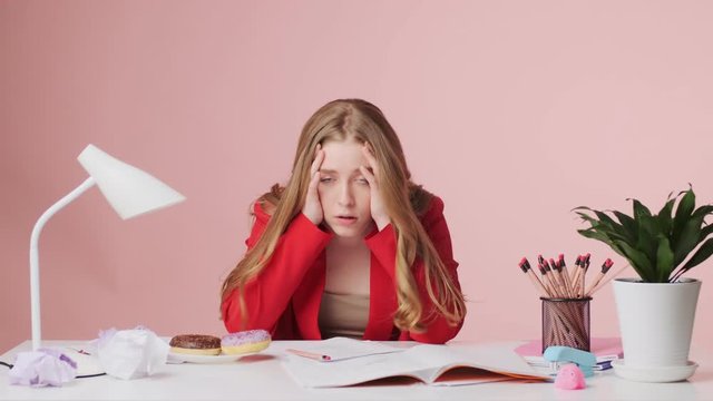 A tired young woman is sitting at the table isolated over pink background