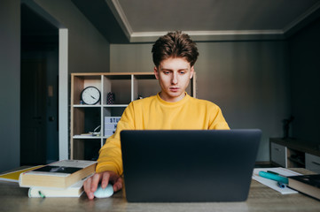 Young male student uses a laptop for distance learning, sits at home with books and notebooks and studies. Guy in the orange sweatshirt is studying to quarantine.
