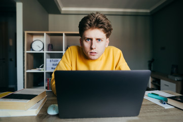Shocked student sitting at home at desk with books and laptop, looking at camera with shocked face. Surprised teen student studying at a distance learning home in a room.