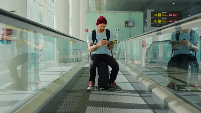 Cool and modern looking young hipster man, sit on suitcase in empty airport terminal. Look at smartphone for online boarding pass or cancelled departures, arrivals flights. Urban nomad travel blogger