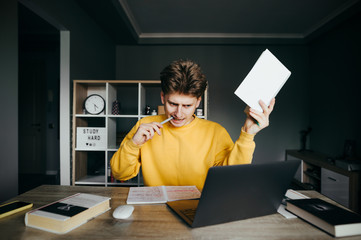 Emotional young man sits at a pulp table with laptop and a book in his hand, looks intently at the...