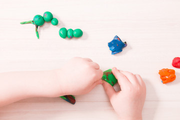 a small child sculpts a caterpillar from green plasticine on a wooden table, developing fine motor skills of hands, playing with a child at home, preparing for kindergarten, stay at home