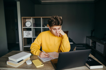 Funny surprised teen student studying at home remotely sitting at a desk with books, writing in a...