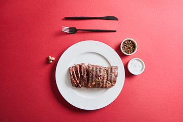 top view of tasty grilled steak served on plate with cutlery and salt and pepper in bowls on red background