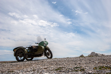 An old Soviet motorcycle with a sidecar stands against the sky with clouds on the stones. Green color. Windshield and round headlight. Horizontal.