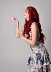 Portrait of a beautiful woman with red hair wearing  a  flowing floral gown.  full length standing pose, isolated against a studio background