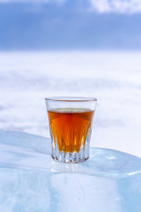 An orange alcoholic drink in a glass stands on an ice piece. Ice melted in the sun. Light blurred background. Copy space. Side view. Vertical.