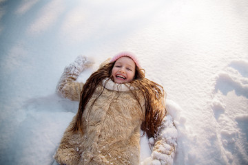 Funny cute girl of 12 years old with curly hair in a pink hat lies with a snowdrift and indulges in snow, showing "Snow Angel"