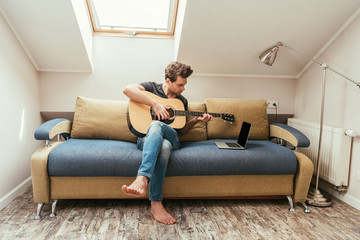 young man playing guitar while sitting on sofa and looking at laptop with blank screen
