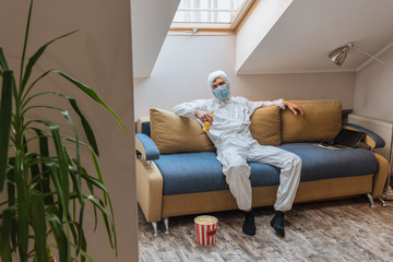 young man in hazmat suit and protective mask holding bottle of beer while sitting on sofa near popcorn bucket on floor