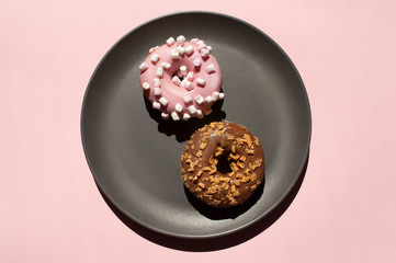 Top view, flat lay of two donats, one decorated with white marshmallows and another chocolate one with caramel-flavored curls on the grey ceramic plate on pink background. Creative art food concept