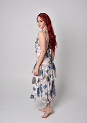Portrait of a beautiful woman with red hair wearing  a  flowing floral gown.  full length standing pose, isolated against a studio background