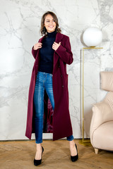 Beauty Fashion Model Girl with curls in burgundy coat in a bright white loft style studio. Beautiful Luxury Winter Woman. Beauty and fashion. Spring and autumn clothes fashion. Girl posing at studio.