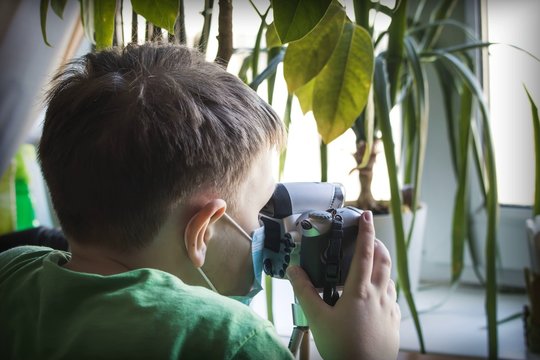 A child in a medical protective mask with a camera in his hands against the background of a window with indoor plants