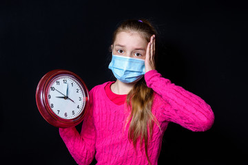 A teenage girl in a pink sweater and a protective mask holds a watch in her hands. On black background.