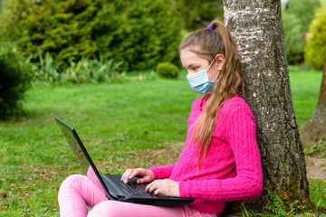 A teenage girl in a pink sweater and protective mask is sitting in nature, leaning against a birch tree, and holding a laptop on her lap