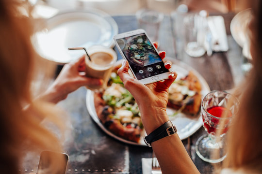 Food, eating, technology, culinary and people concept - woman hands with smartphone photographing pizza with girlfriend at restaurant  