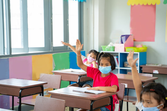 Group of Asian children wearing protective mask to Protect Against Covid-19 with teacher sitting in classroom and raising hands at desk in classroom,Social Distancing.