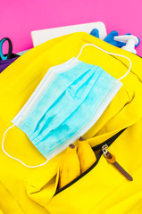 Yellow backpack full of school must-have tools also with face mask, hydro alcoholic gel and latex gloves to protect from covid-19 on the pink background