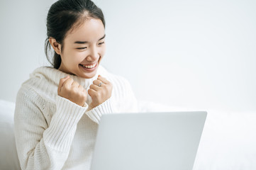 A woman wearing a white shirt on the bed and playing laptop happily.