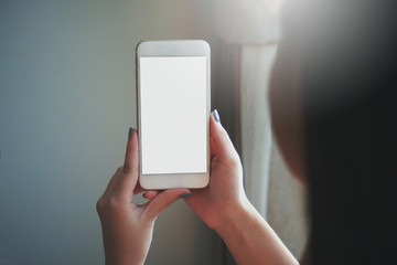 Closed up image taken from the back of a woman holding and using a white blank screen phone. White flare from the window.