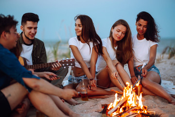 Group of young friends sitting on beach and fry sausages. One man is playing guitar. Summer holidays, vacation, relax and lifestyle consept. Camping time.