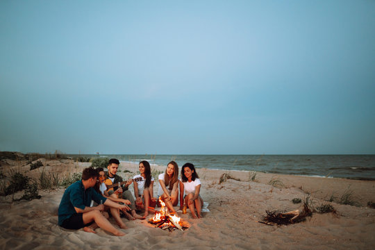 Group of young friends sitting on beach and fry sausages. One man is playing guitar. Summer holidays, vacation, relax and lifestyle consept. Camping time.