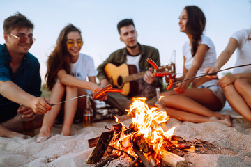 Fried sausages on bonfire. Group of young friends sitting on beach and fry sausages. Summer holidays, vacation, relax and lifestyle consept. Camping time. - Powered by Adobe