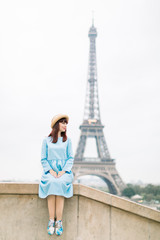 Happy young woman in romantic blue dress and hat posing near the Eiffel tower in Paris, sitting on the stairs at Trocadero place and looking aside