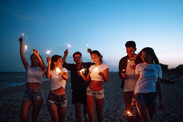 Group of friends at night on the beach with sparklers. Young friends enjoying on beach holiday. Summer holidays, vacation, relax and lifestyle concept. - Powered by Adobe