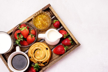 Fototapeta na wymiar Wooden Tray with Breakfast Freshly Baked Buns with Raisins and Cinnamon Cup of Black Coffee Cream Ripe Strawberry and Orange Jam in Glass Bowl