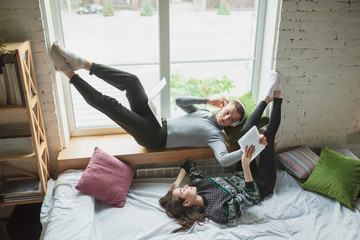 Using devices lying down in bedroom near window. Quarantine lockdown, stay home concept - young beautiful caucasian couple enjoying new lifestyle during insulation. Happiness, togetherness, healthcare