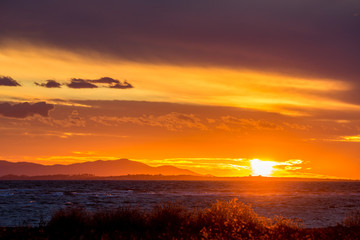 Sun is setting down to the West partially hidden behind curly clouds near the fishermen village of Porto Lagos, Northern Greece. Mountain range and sea water in darkness. Blurred bushes in foreground