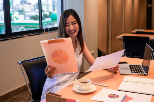 Asian businesswoman in white dress looking at report chart happily with smile at her desk with laptop, coffee cup and notebook. Business concept for work from or success woman