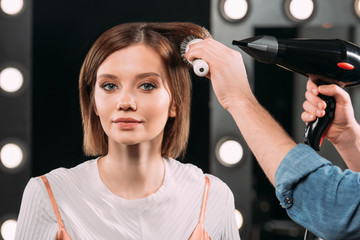 Makeup artist doing hairstyle with hairbrush and hair dryer to attractive woman