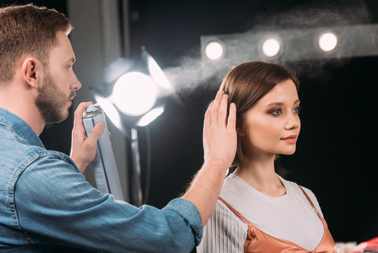 Handsome makeup artist using hair spray while doing hairstyle to beautiful model in photo studio