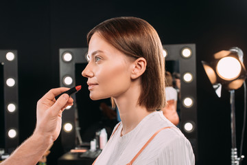 Side view of makeup artist holding red lipstick while doing makeup to beautiful model in photo studio