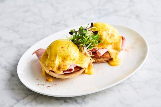 Eggs Benedict on a white plate, side view. Side view on eggs Benedict with arugula, bacon and hollandaise sauce on gray wooden background.
