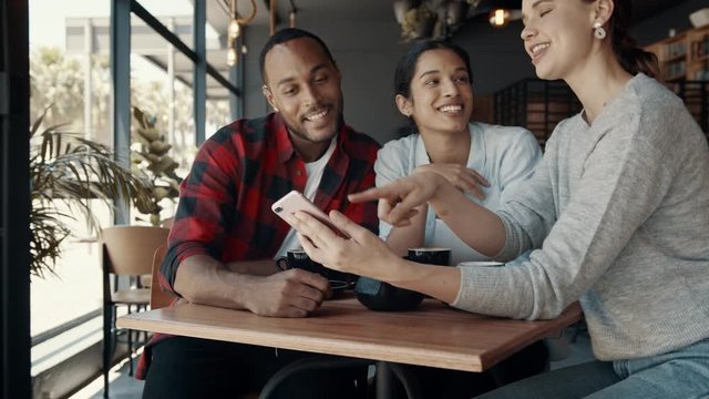 Group of man and women sitting in a cafe looking at a mobile phone and laughing. Best friends hanging out at a coffee shop.
