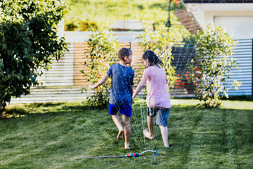 Young boy and girl playing with the garden sprinkler. Summertime activities at home during isolation.
