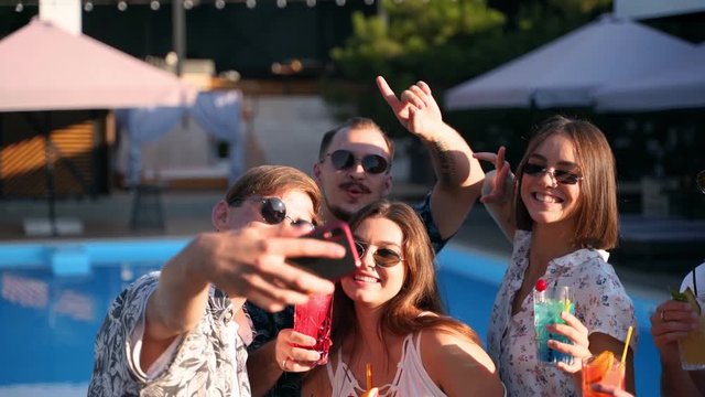 Smiling friends taking selfie with smartphone at poolside party with fresh colorful cocktails standing by swimming pool on sunny summer day. Woman taking photo at luxury villa on tropical vacation.