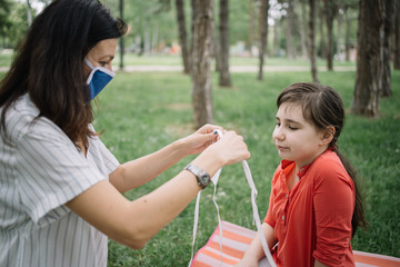 Obraz premium Mother with mask putting mask to her daughter outdoor. Woman wearing medical mask setting mask to her kid while sitting in park.