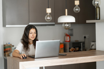 Beautiful young brunette girl working on a laptop at home in the kitchen.