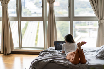 A beautiful young brunette girl is working on the laptop while lying on a bed by a panoramic window with a beautiful view from a high floor. Stylish modern interior.Shopping on the Internet
