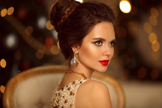 Luxurious portrait of elegant woman with wedding hairstyle and makeup. Beautiful brunette girl with golden jewelry in prom dress sitting on modern chair over bokeh lights xmas decorations.