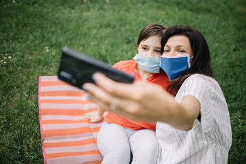 Happy family with masks sitting on mat in park and taking selfie. Mom and kid wearing medical masks while lying in park and taking photos with phone.