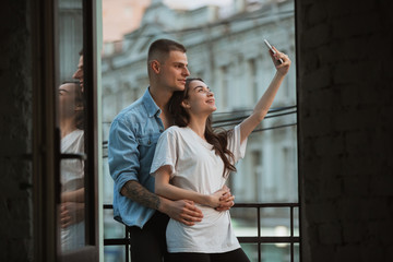 Standing on the balcony, hugging, selfie. Quarantine lockdown, stay home concept - young beautiful caucasian couple enjoying new lifestyle during coronavirus. Happiness, togetherness, healthcare.