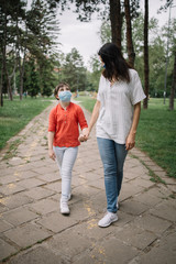 Fototapeta premium Woman holding little girl's hand while walking in park. Mom and child walking on park alley while wearing antivirus masks during coronavirus pandemic.