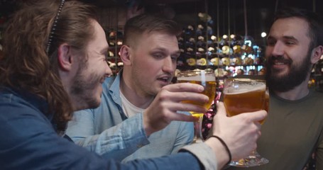 Caucasian male friends cheering up in bar when watching sport game. Handsome men doing cheers gesture with glasses of beer in pub. Football fans watch match on TV and drinking.