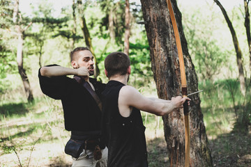 A man teaches to shoot with a bow. Two men, one teacher. Archery in nature. The boy holds a bow in his hands.
